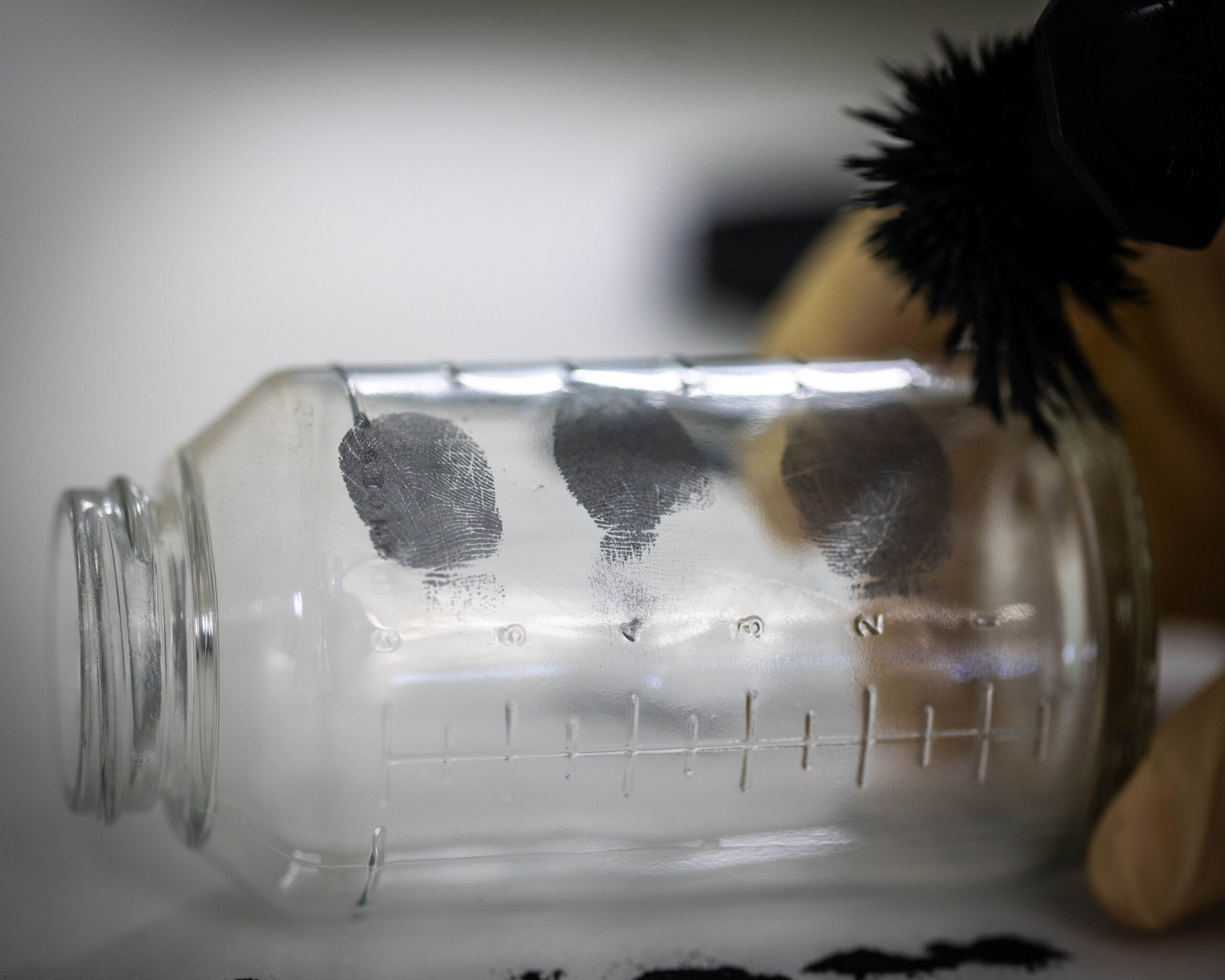 A glass jar with three fingerprints covered with dust and a brush is shown